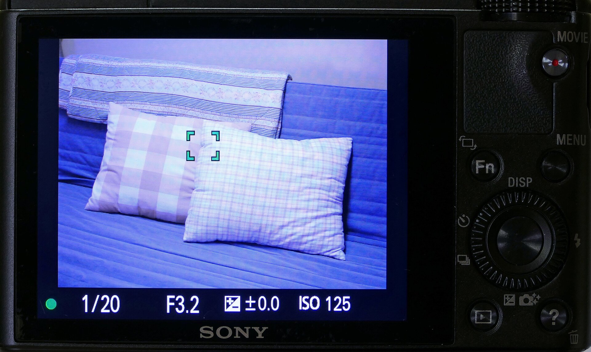 Zdjęcie przedstawia widok wyświetlacza aparatu fotograficznego, na którym widoczne jest wykonane zdjęcie (wersalka przykryta niebieską kapą z 2 poduszkami). Poniżej widoczne są parametry ustawienia autofocusa. Po lprawej stronie panel z pokrętłami i przyciskami aparatu fotograficznego.