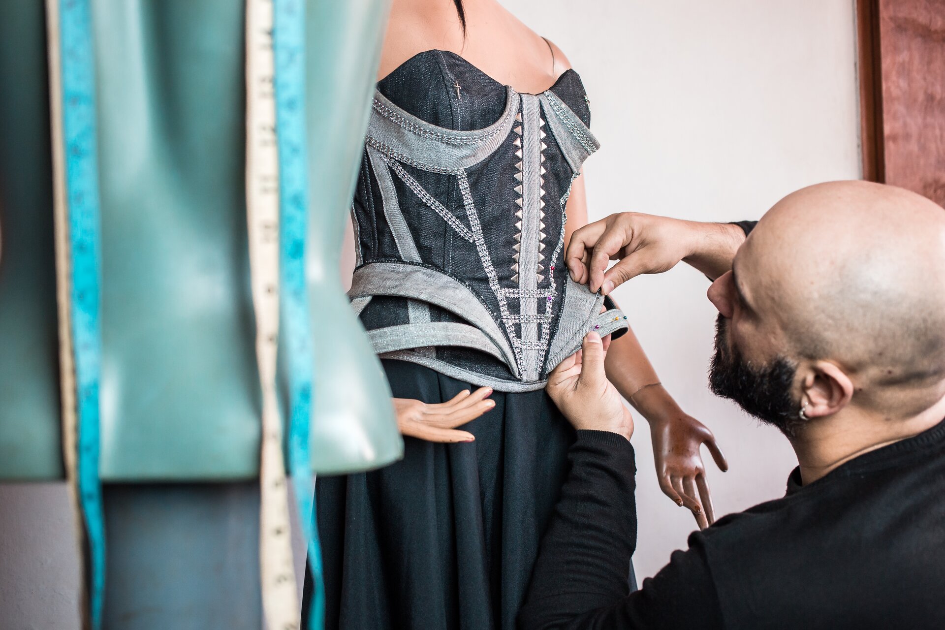 Ilustracja przedstawia łysego mężczyznę projektującego damską sukienkę na manekinie. Sukienka jest czarno‑szara. Po lewej stronie znajduję się miarka krawiecka.