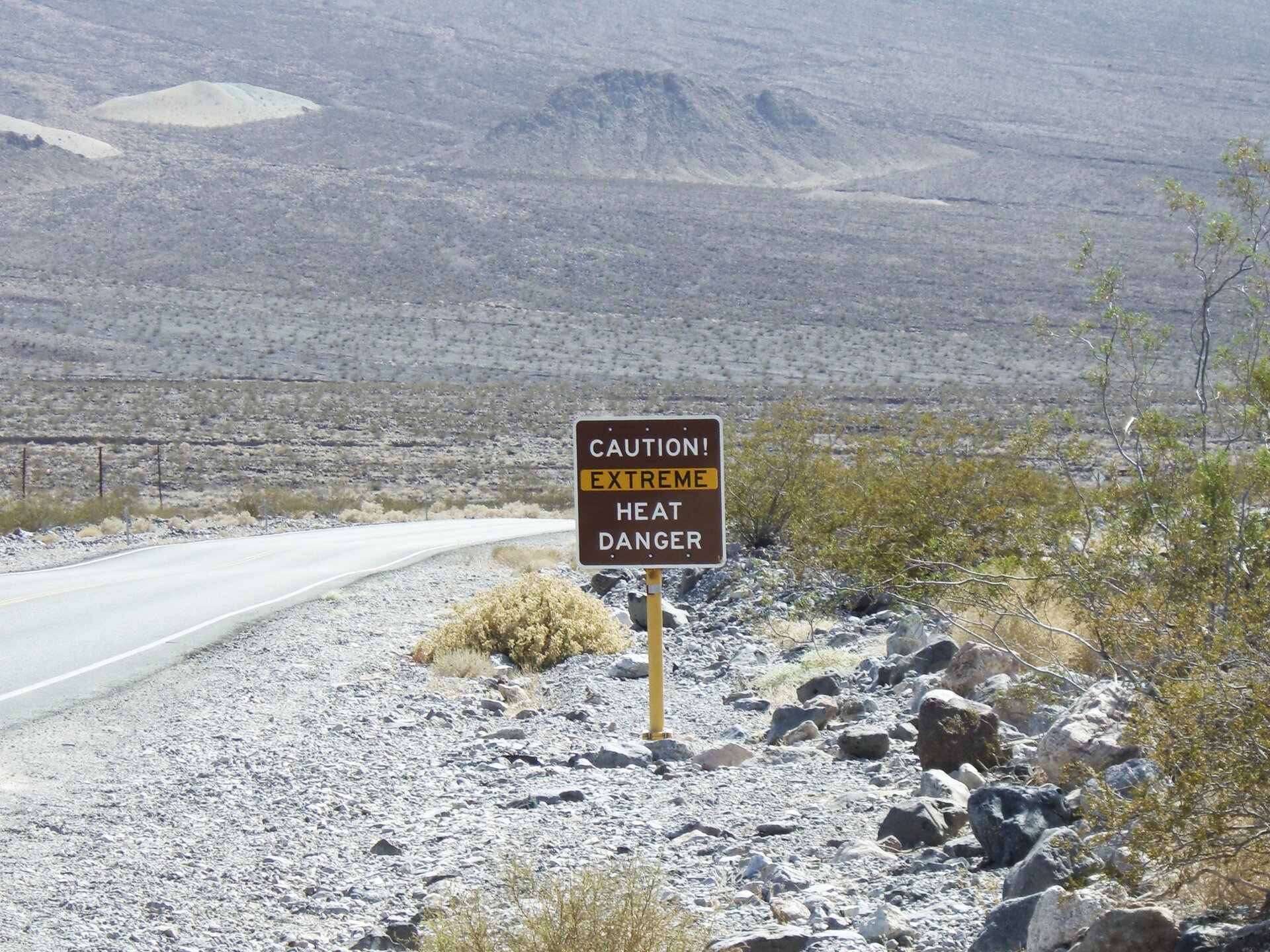 Fotografia przedstawia tablicę informująca o zagrożeniu nadzwyczaj wysoką temperaturą powietrza. Tablica jest prostokątna, brązowa, z białymi i zaznaczonymi na żółto napisami w języku angielskim. Stoi na skraju Doliny Śmierci na pustyni Mojave. Przy żółtym słupie tablicy leżą szare i brązowe kamienie, między którymi rosną kępy roślin – traw i niskich krzewów. W tle łańcuchy górskie.