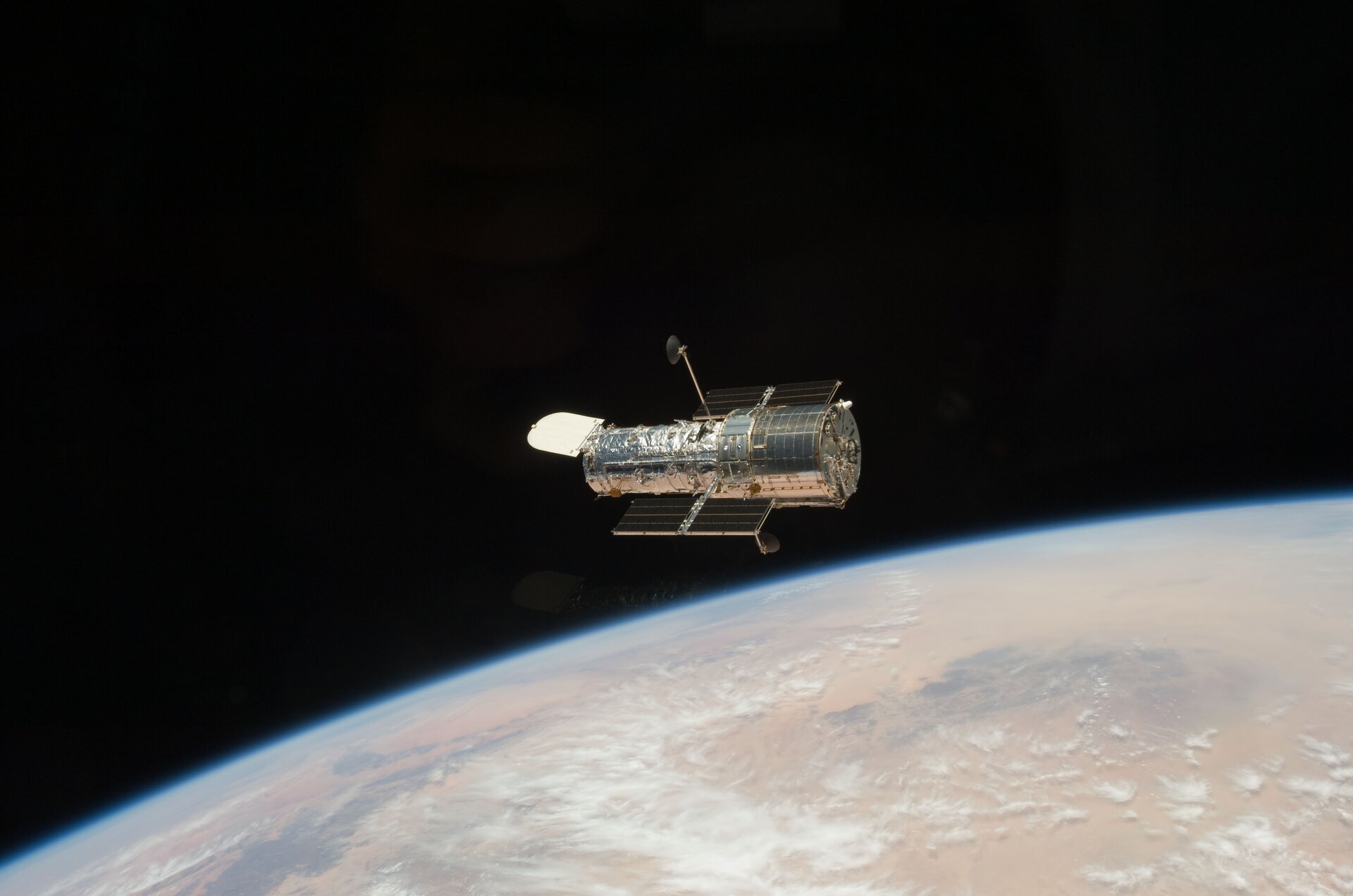 Zdjęcie przedstawia Kosmiczny Teleskop Hubble'a w przestrzeni kosmicznej. Teleskop ma w przybliżeniu kształt srebrnego walca, jest ułożony poziomo. U góry i u dołu tego walca widoczne są czarne panele słoneczne. W tle widoczne są fragment Ziemi i czerń kosmosu.