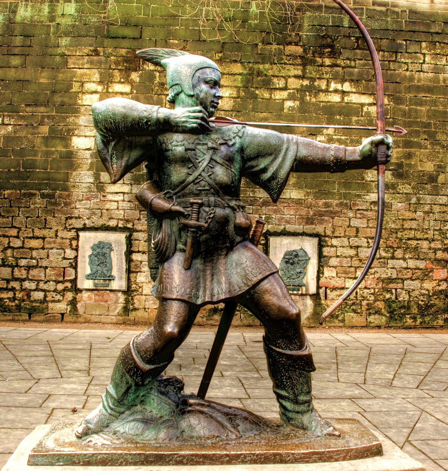 Zdjęcie przedstawia pomnik. To strzelający z łuku mężczyzna w krótkiej szacie do połowy uda. Mężczyzna ma długie buty sięgające do kolan, a na głowie czapkę z piórem. U boku przypięty sztylet i róg.