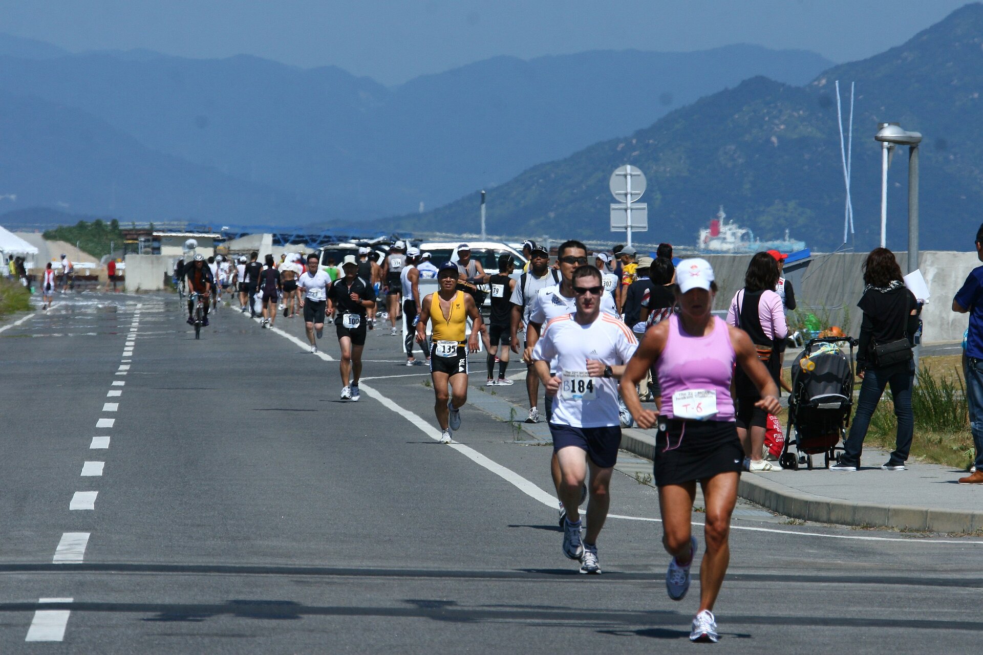 Kolorowe zdjęcie przedstawia biegnących jezdnią maratończyków. Mężczyźni i kobiety w koszulkach z krótkim rękawkiem lub na ramiączkach i krótkich spodenkach. Każda osoba ma przypięty na brzuchu numer startowy. W tle widać wysokie góry.