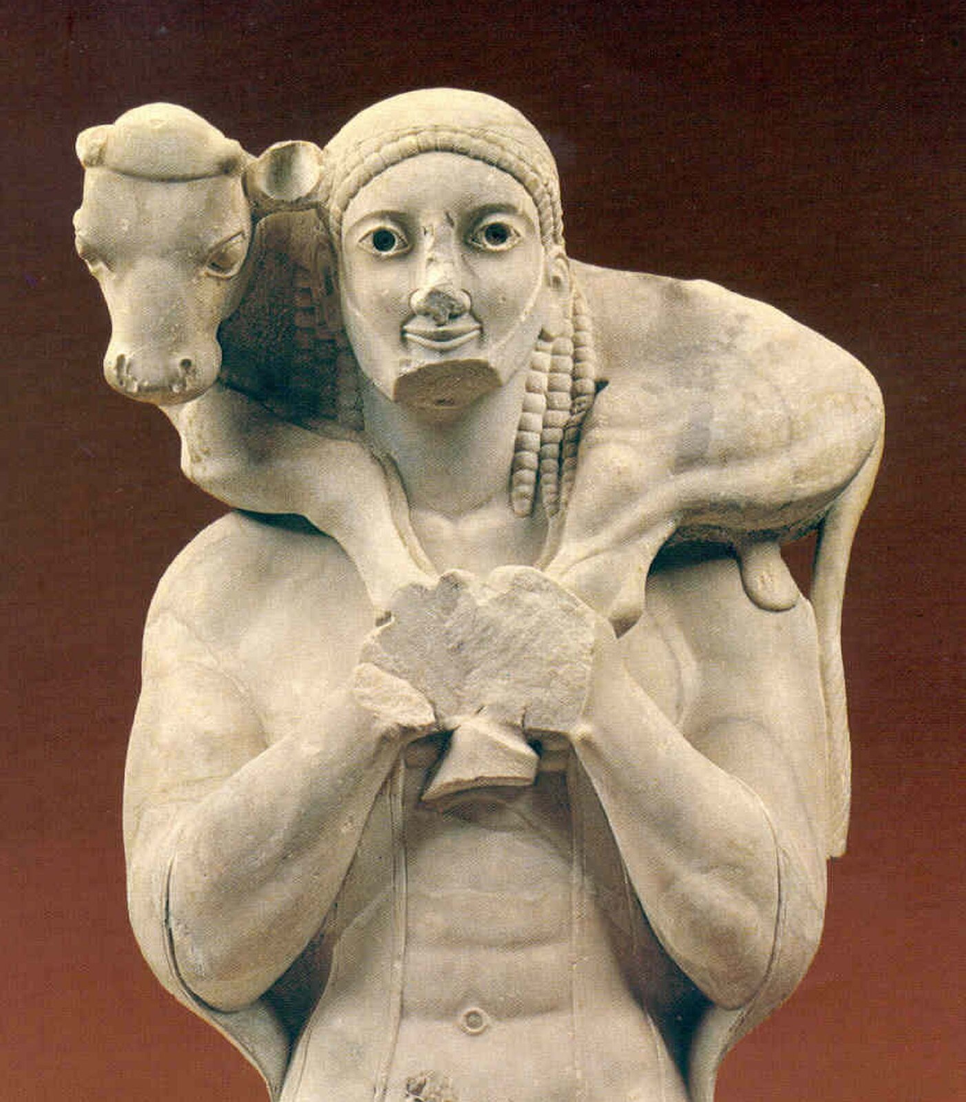 Ilustracja przedstawia amforę grecką. Na zdjęciu widnieje wysmukłe, gliniane naczynie o pękatym brzuścu i wydłużonej szyjce, przykrytej pokrywką z uchwytem. Posiada jeden wydłużony, smukły uchwyt. Amfora ozdobiona jest drobnym, czerwono-brązowym ornamentem geometrycznym i utrzymana jest w jasnej tonacji.