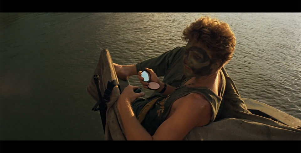 Kadr przedstawia mężczyznę, który płynie łodzią po rzece. W ręku trzyma puderniczkę z lusterkiem. Mężczyzna ma twarz pomalowaną w ciemnozielone plamy. Spogląda w bok. Ubrany jest w ciemnozieloną koszulkę na ramiączka i spodnie.