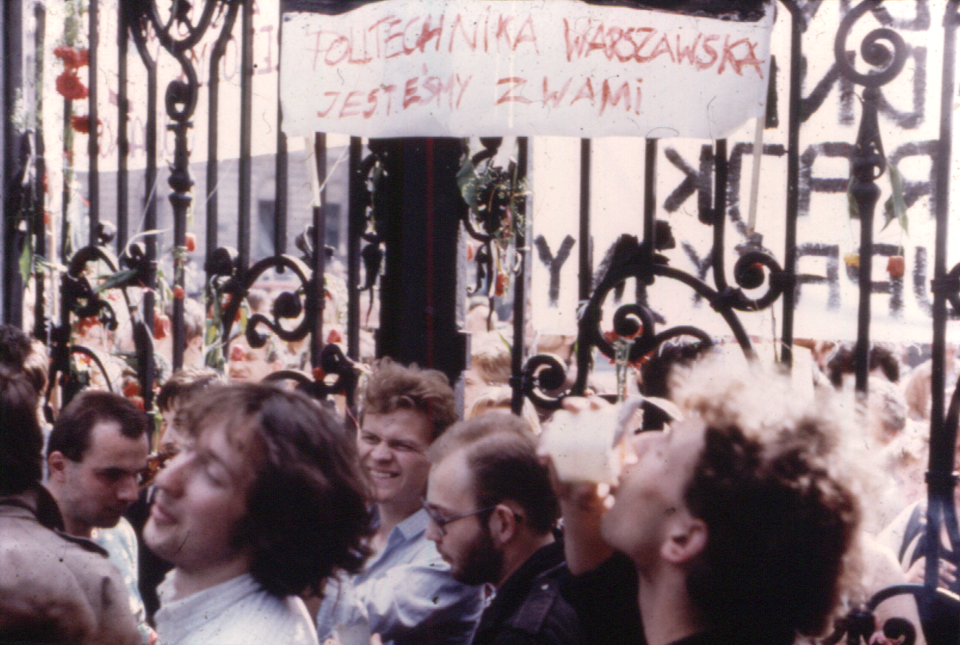 Zdjęcie przedstawia dużą grupę młodych ludzi. Stoją oni przy żelaznej, kutej bramie. Wisi na niej transparent z napisem: Politechnika Warszawska jesteśmy z Wami.