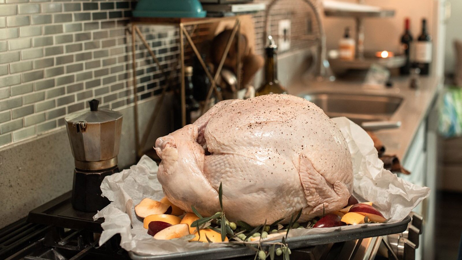 Zdjęcie przedstawia przygotowanego do pieczenia indyka leżącego na blaszce. Na skórze indyka widoczne są przyprawy. Indyk położony jest na warstwie połówek jabłek i gałązkach rozmarynu. Poniżej znajduje się pergamin do pieczenia. W tle widoczny jest blat kuchenny ze zlewem i wyposażeniem.