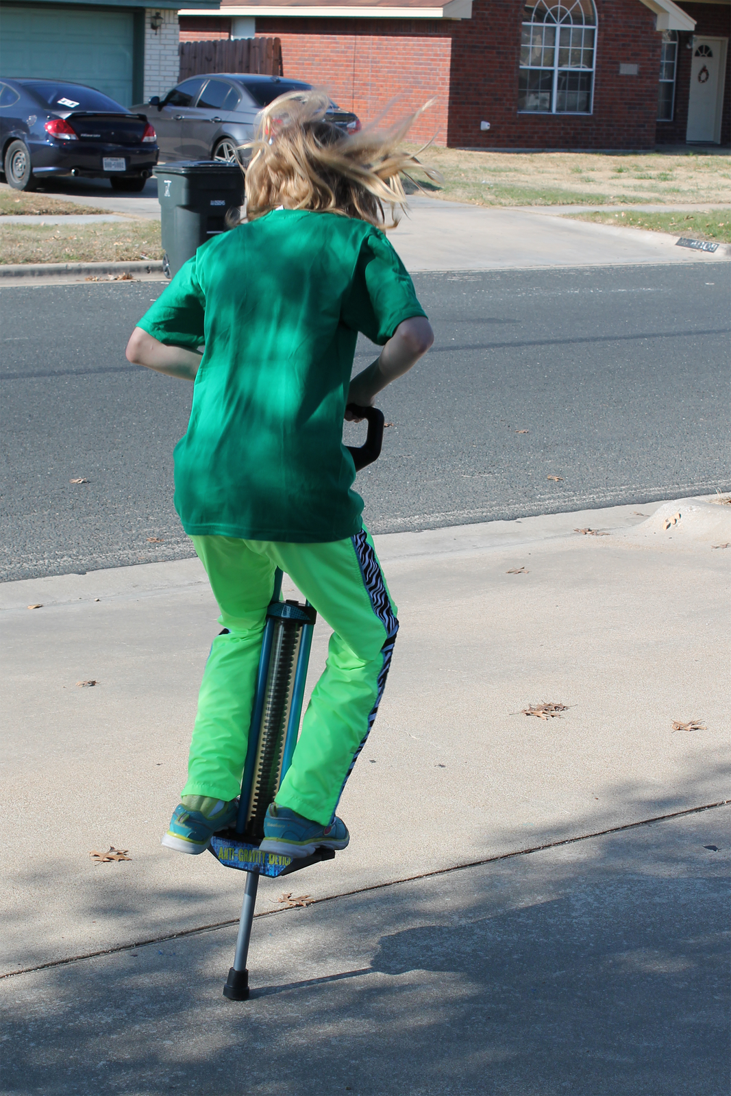 Fotografia przedstawia dziecko skaczące na zabawce pogo. Zabawka ta składa się z rurki, na którą nałożona jest sprężyna oraz platforma do stania oraz przedłużenie mające służyć do trzymania w trakcie skakania na urządzeniu. Dziecko widoczne jest od tyłu.