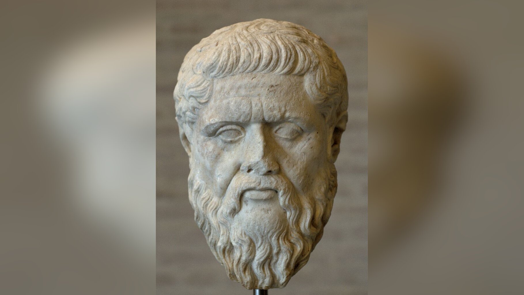 Zdjęcie przedstawia rzeźbę głowy mężczyzny. Mężczyzna jest dojrzały. Jego twarz jest owalna, oczy głęboko osadzone, czoło wysokie. Mężczyzna ma krótkie włosy, gęstą brodę i wąsy. Rzeźba ma lekko utrącony nos.