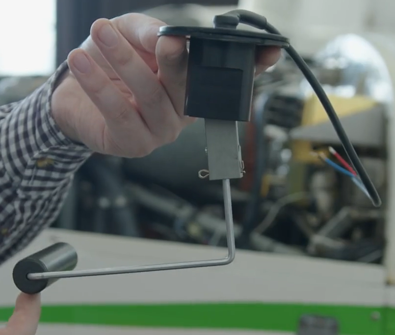 Zdjęcie przedstawia paliwomierz pływakowy. Widoczny jest trzymany w ręce paliwomierz pływakowy. W górnej części urządzenia znajduje się kabel połączony z wieczkiem, pod którym zawieszony jest prostopadłościan. Do jego dolnej podstawy zamontowany jest metalowy pręt, który następnie zgina się pod kątem prostym poziomo w lewo. Na końcu pręta znajduje się cylinder zamocowany na pręcie jedną z podstaw i leżący poziomo, pod kątem prostym do pręta.