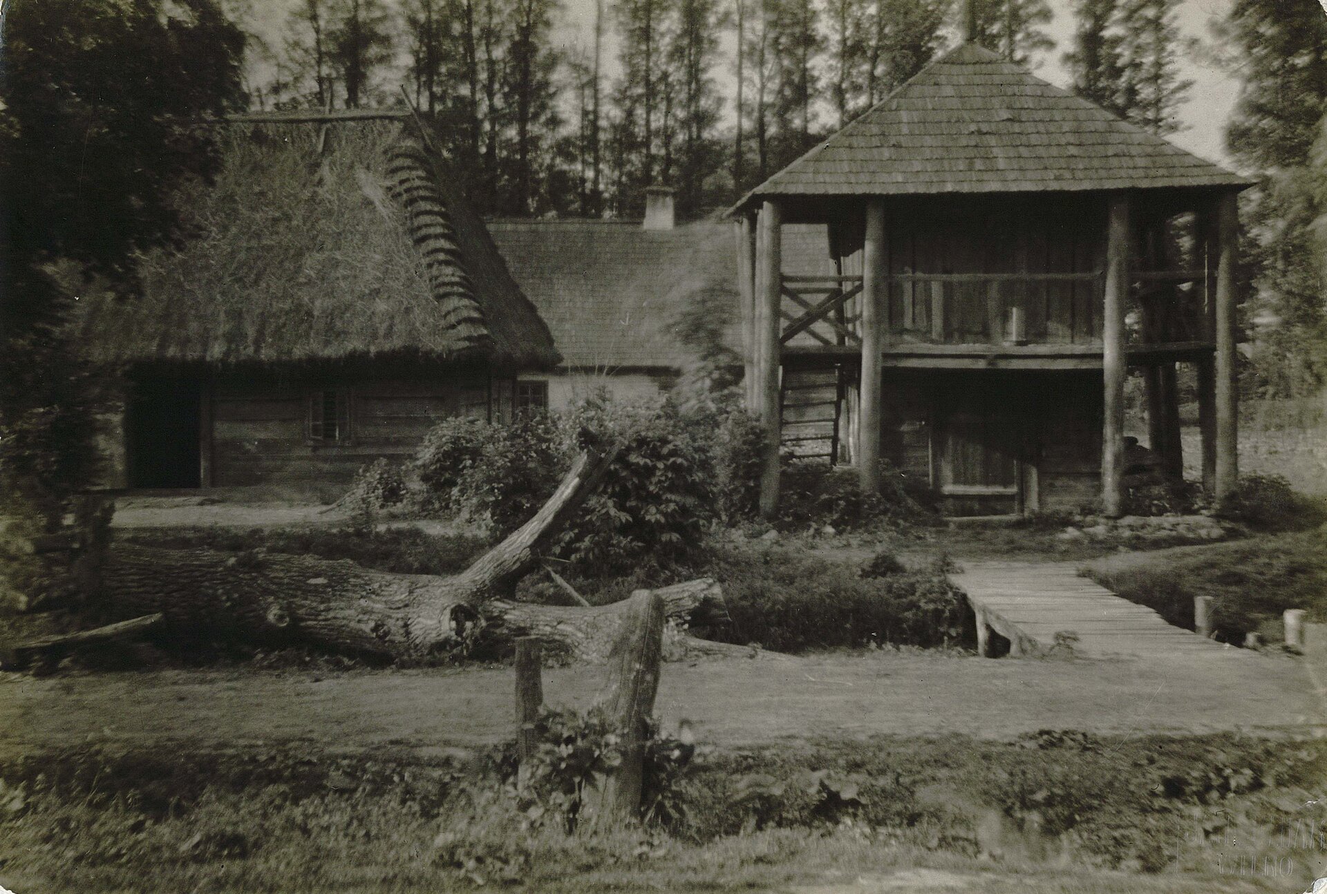 Stare czaro‑białe zdjęcie przedstawiające starą drewnianą chatę z zagrodą. Od frontu zagrody przewrócone drzewo. Po prawej stronie drewniany, dwupiętrowy budynek. Po lewej stronie duża drewniana chata ze strzechą. W oddali budyek. 