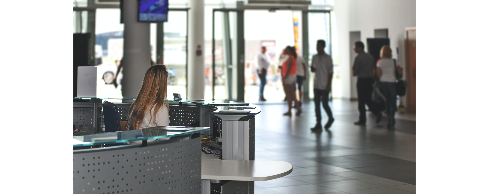 Zdjęcie przedstawia stanowisko kasy biletowej na lotnisku. Za ladą w kasie siedzi kobieta z długimi włosami. W tle widoczni są ludzie.