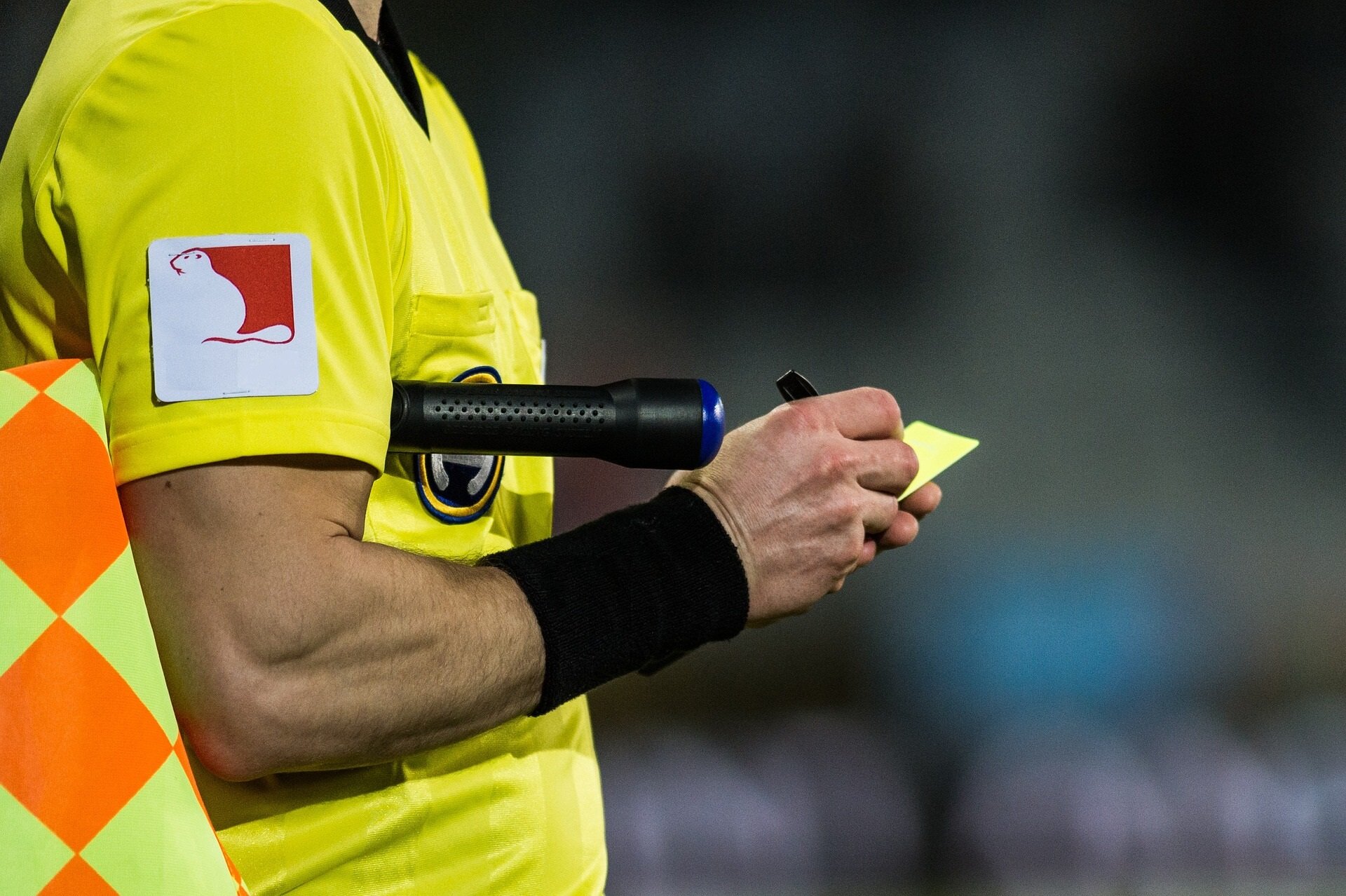 Zdjęcie przedstawia sędziego piłkarskiego, który pod pachą trzyma chorągiewkę w żółto‑pomarańczową kratę i zapisuje coś na żółtej kartce.