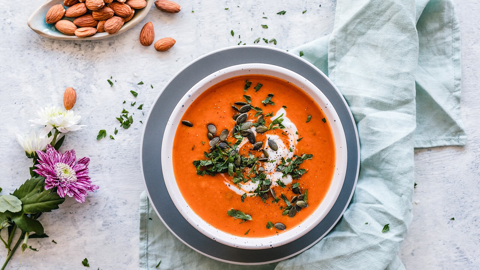 Zdjęcie przedstawia postawiony na stole talerz, w której widoczna jest zupa w kolorze pomarańczowym przyozdobiona pestkami dyni. Obok widoczne pestki migdałów oraz trzy kwiaty.  