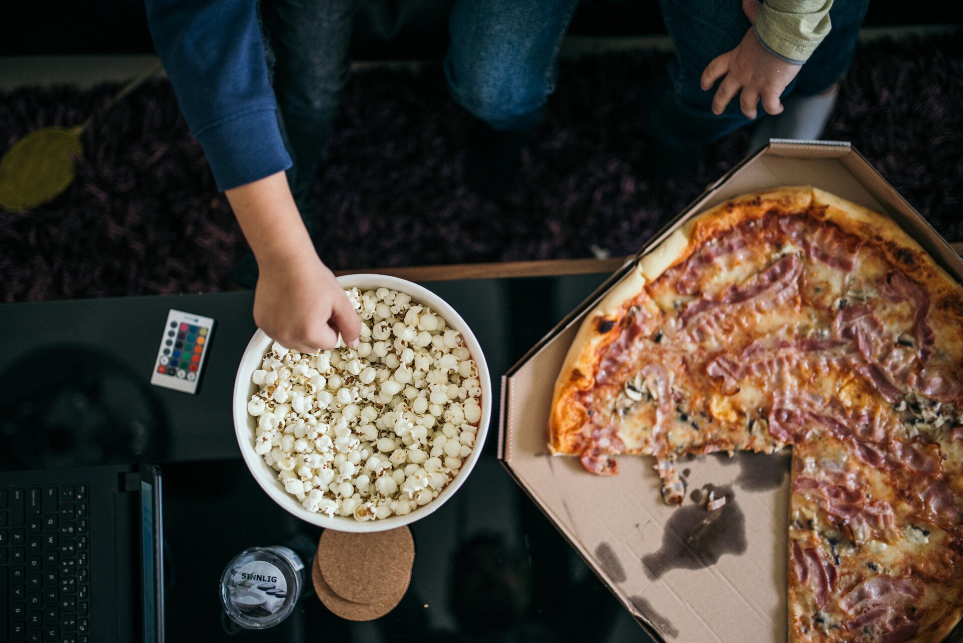 Zdjęcie przedstawia ukazany z góry blat stołu. Leży na nim w duża pizza w kartonie, stoi miska z popcornem, do której sięga ręka mężczyzny. Nieopodal jedzenia, na stole stoi laptop.  