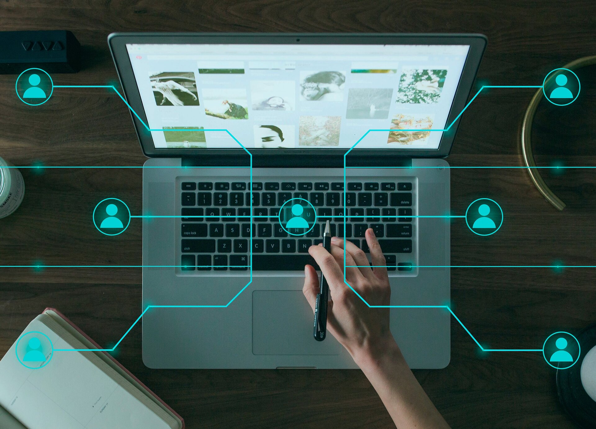 Ilustracja przedstawia zdjęcie otwartego laptopa stojącego na drewnianym blacie oraz prawej dłoni trzymającej długopis i dotykającej klawiatury laptopa. Na monitorze laptopa widoczne są różne zdjęcia z opisami. Na zdjęcie nałożona została grafika przedstawiająca siatkę połączeń, złożona z niebieskich linii, na końcu których znajdują się okręgi z wpisaną ikonką człowieka. Pośrodku klawiatury widoczna jest ikonka, od której w obie strony odchodzi siatka połączeń.