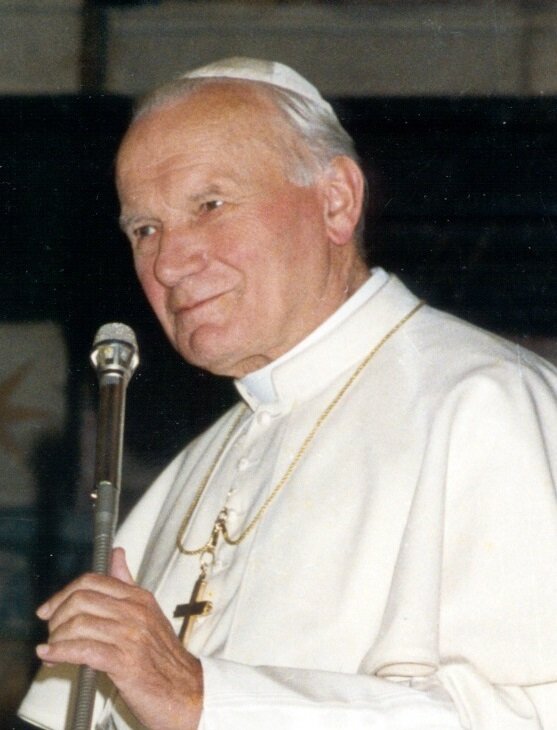 Zdjęcie portretowe Jana Pawła Drugiego. Ubrany jest w biały strój papieski, na piersi ma złoty krzyż. Uśmiecha się, w lewej ręce trzyma mikrofon.
