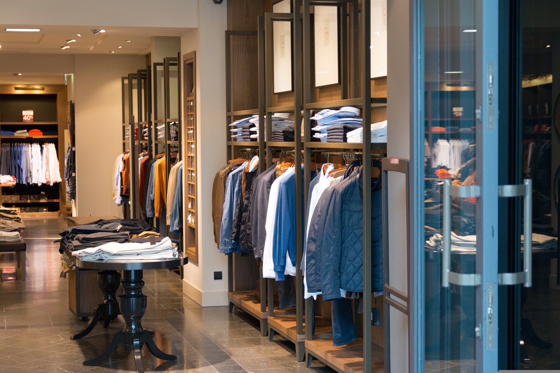 Grafika przedstawia sklep odzieżowy. Widoczne ubrania na wieszakach, półkach i stołach.