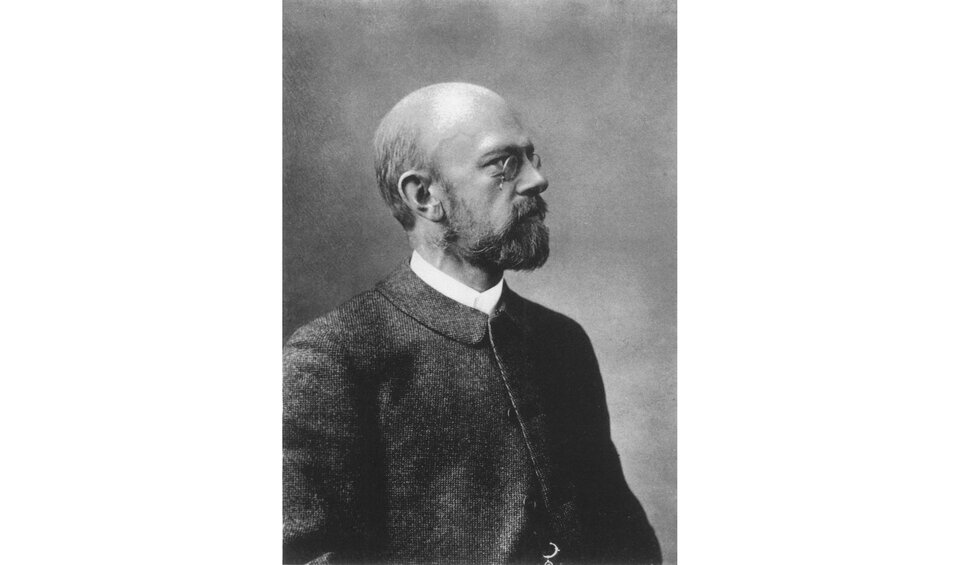 Ilustracja prezentuje mężczyznę w okularach, z brodą i wąsami. Mężczyzna zdaje się patrzeć w dal.