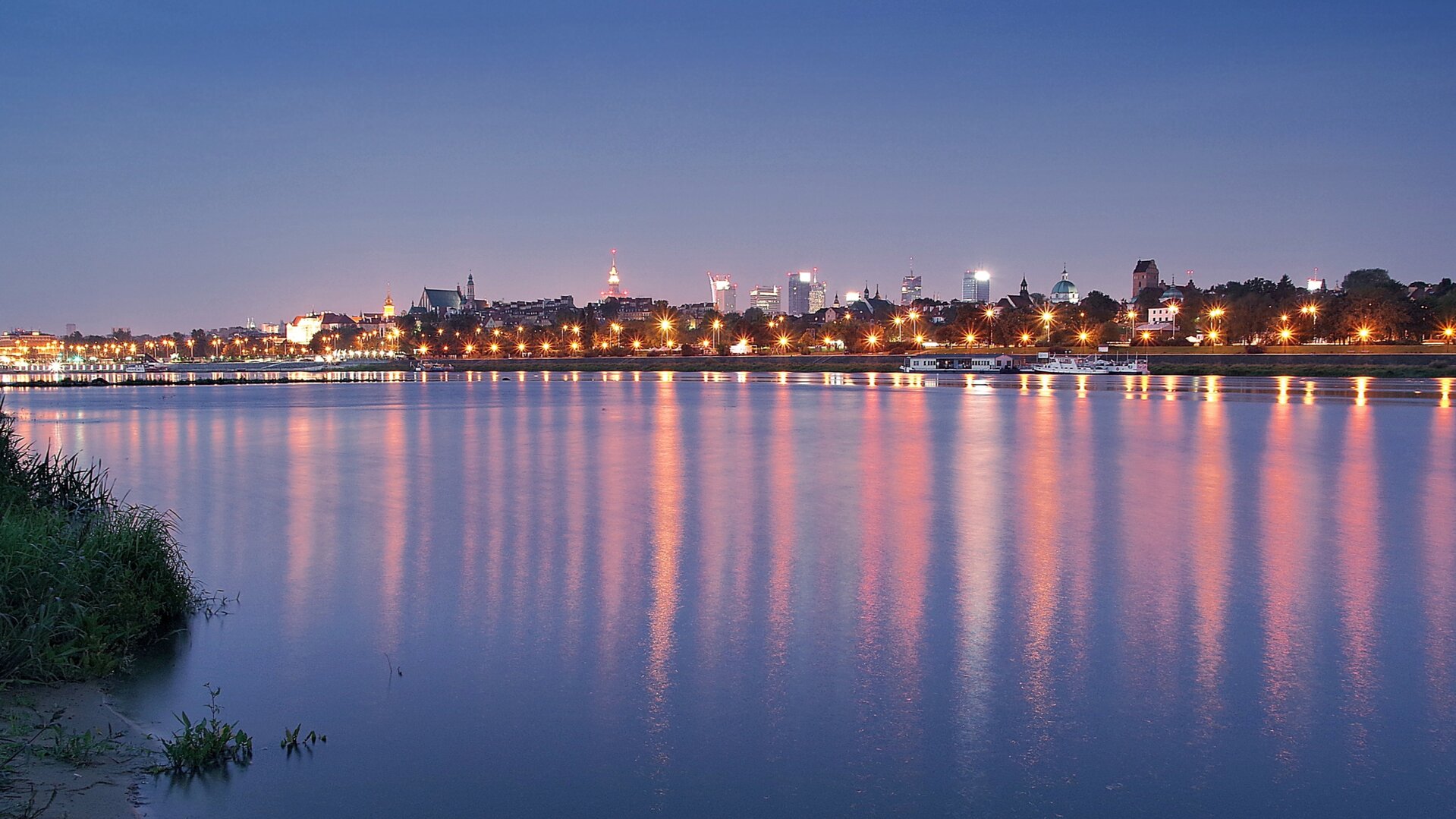 Fotografia przedstawiająca widok z brzegu Wisły na panoramę Warszawy. Zdjęcie zrobione o zmroku. W wodzie odbijają się różnobarwne refleksy świetlne. W tle oświetlony brzeg z panoramą miasta.