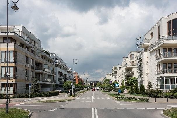 Zdjęcie przedstawia szeroką ulicę. Po jej obu stronach znajdują się czteropiętrowe budynki. Dookoła rosną krzewy. 