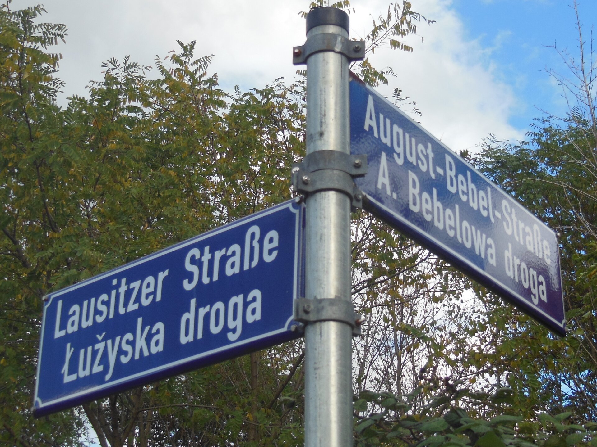 Na zdjęciu słup uliczny z nazwami ulic. Niebieskie tabliczki, białe napisy – niemieckie i łużyckie nazwy ulic.
