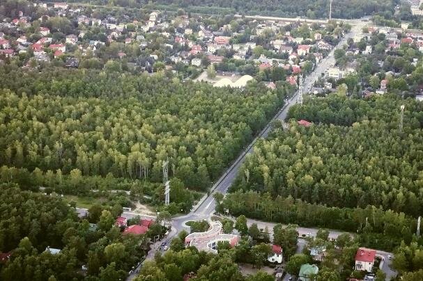 Zdjęcie przedstawia widok z góry na dzielnicę Warszawy. Na pierwszym planie znajduje się las, przedzielony pasmem drogi. W tle osiedle domków jednorodzinnych. 